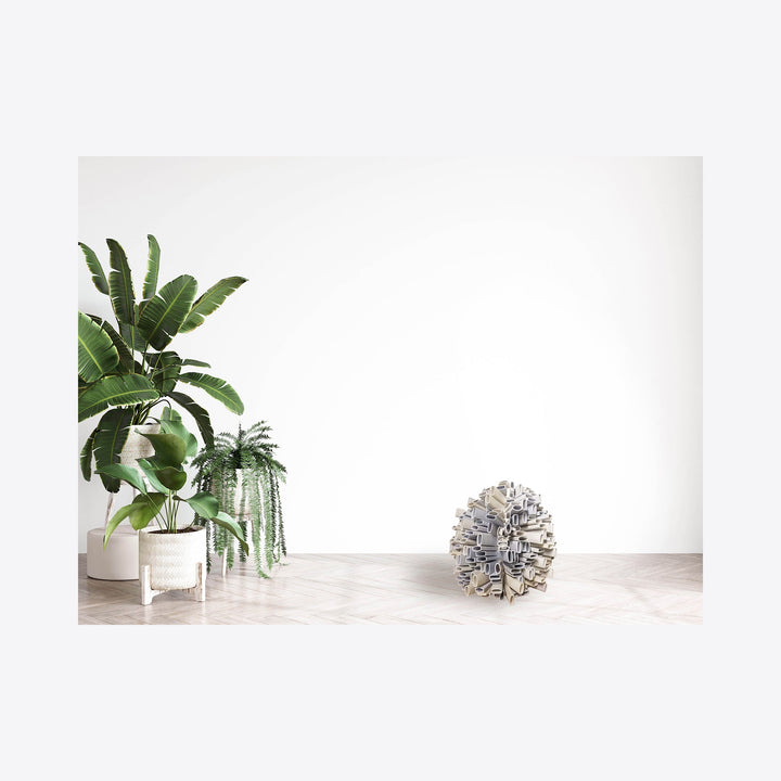 The artwork 'Implosion (Behind the complaint)' by Kate Rusek features a spherical sculpture crafted from reclaimed plastic, steel, and copper, positioned on a light-colored floor against a white wall. The sculpture contrasts with lush green tropical plants in white planters to the left, creating a minimalist, contemporary aesthetic.