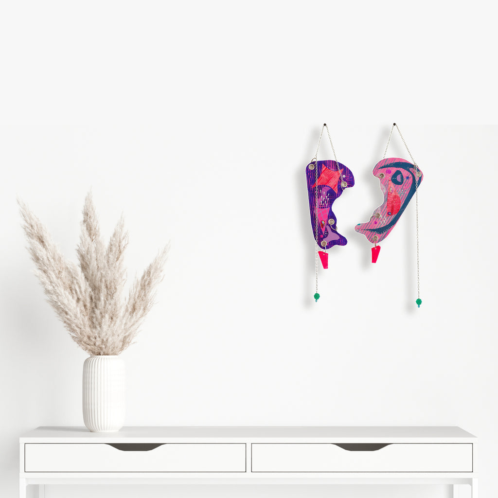 The artwork 'Rise Above' by Sunny Chapman features two hanging abstract face profiles in vibrant purple and pink hues, adorned with glitter, jewels, and metallic accents. The mixed-media pieces are suspended by chains and grommets, with delicate beaded details and turquoise accents, creating a whimsical wall decoration against a clean white background. - Sunny Chapman