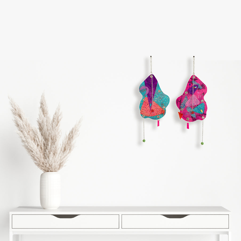 Artwork 'The Day the World Turned Glitter' features whimsical abstract shapes in vibrant turquoise and magenta, adorned with dangling elements. Mixed media wall pieces combine glitter, jewels, and metallic accents creating dynamic textural contrast. Displayed alongside minimalist white furniture and pampas grass. By Sunny Chapman.