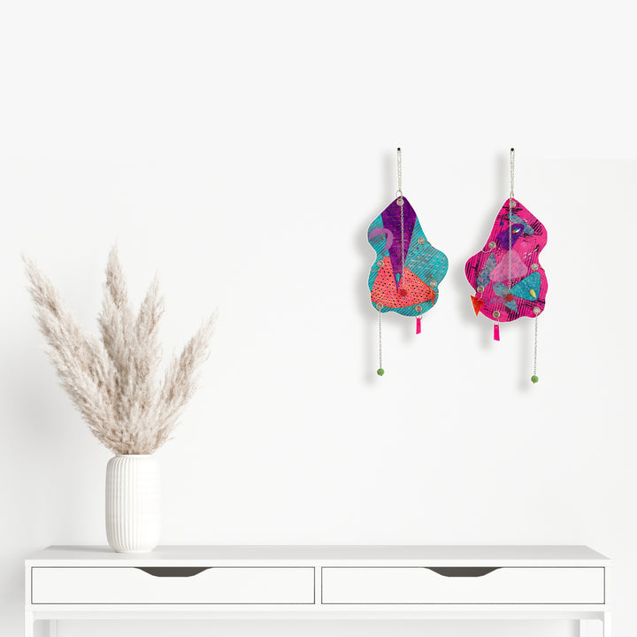 Artwork 'The Day the World Turned Glitter' features whimsical abstract shapes in vibrant turquoise and magenta, adorned with dangling elements. Mixed media wall pieces combine glitter, jewels, and metallic accents creating dynamic textural contrast. Displayed alongside minimalist white furniture and pampas grass. By Sunny Chapman.