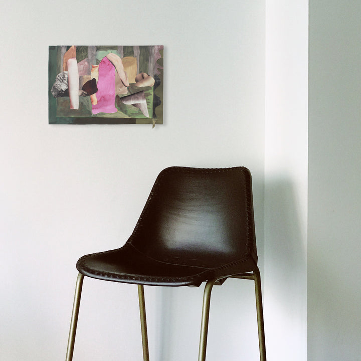The artwork 'In The Stillness' by Winnie Sidharta features abstract watercolor composition with pink and gray geometric shapes layered against muted earth tones. The 12x18 inch piece showcases delicate painted cutouts and fluid watercolor techniques, creating a serene, contemporary aesthetic on paper. Wall art displayed above modern brown leather chair.