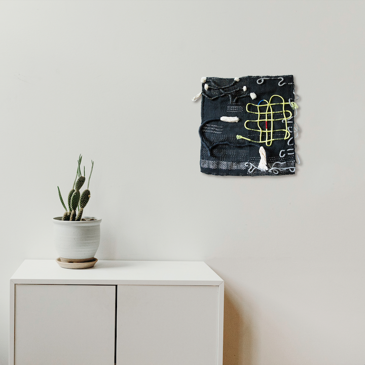 The artwork 'Intervened Couplets' by Kathie Halfin features a contemporary textile composition with black fabric base interwoven with neon yellow flux yarn creating geometric patterns. Silver floss and nylon rope add textural contrast against white wall. Minimalist display above white cabinet with succulent plant.