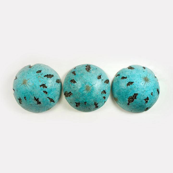 The artwork 'IS' by Nora Chavooshian features three spherical forms in vibrant turquoise cast forton with bronze patina. Each sphere displays delicate star-burst patterns and irregular dark markings scattered across their surfaces, creating a celestial or cosmic effect. The spheres are arranged horizontally, measuring 74 inches in width and 24 inches in height.