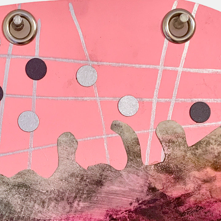 The artwork 'Juggler' by Sunny Chapman features an abstract composition on pink paper with intersecting silver lines creating a grid pattern. Black and metallic circular elements float above a textured brown-grey form, suggesting movement and balance. Metal grommets anchor the top corners of this mixed media piece combining monotype prints and marker details.