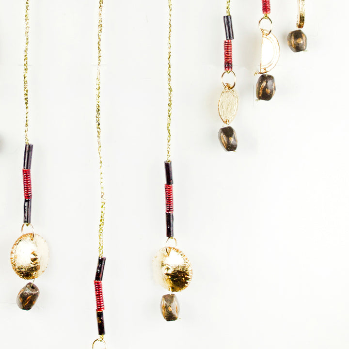 The artwork 'June' by Adina Andrus features delicate hanging pendants with brown smoky quartz beads suspended from golden chains. Each pendant combines red-wrapped metal segments, 24k gold-glazed stoneware elements, and translucent stones, creating an elegant vertical composition against a white background.