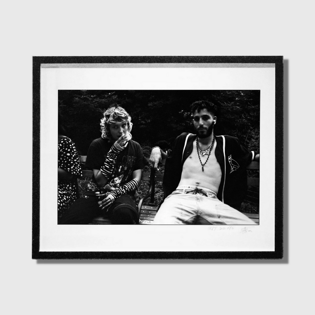 The artwork 'Kids In Washington Square Park' by Mari Sarai captures a candid black and white moment in an urban park setting. A monochromatic composition featuring two figures resting on wooden benches amid foliage. The image showcases contrasting textures between sharp architectural elements and soft natural backgrounds, professionally framed in classic black. Edition 20, 40x32 inches, by Mari Sarai.