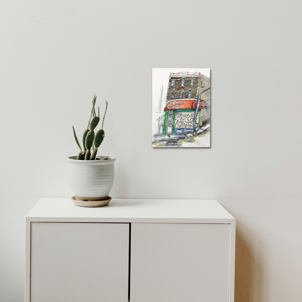 The artwork 'La Villita Bakery' by Andrew Chan features a watercolor and ink illustration of a charming storefront with distinctive architectural details. The piece combines loose brushstrokes and detailed linework, depicting a colorful urban scene with green and red signage. The artwork hangs above a minimalist white cabinet beside a potted cactus, shown in a lifestyle setting.