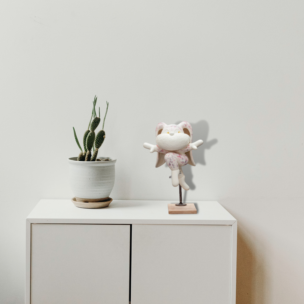 The artwork 'Leap of Faith' by Yusuke Ochiai features a whimsical white resin sculpture of a cheerful cat figurine balanced on a wooden base. The cat wears a delicate pink-patterned dress and spreads its arms wide in a joyful pose, enhanced with 22k gold leaf accents against a textured grey background. The sculpture embodies playful movement and artistic grace.