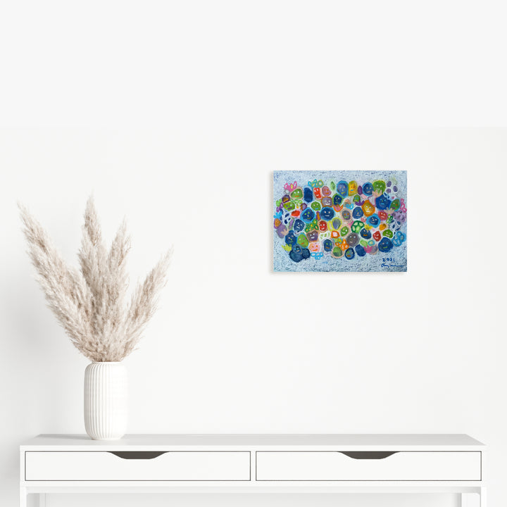The artwork 'Living in a Community' by Ayane Kurai features vibrant circular patterns in blue, green, and multicolored dots scattered across a light blue watercolor background. The whimsical composition creates an abstract, bubble-like effect, expertly rendered in watercolor and cray-pas on paper, measuring 11 x 14.5 inches. By Ayane Kurai.