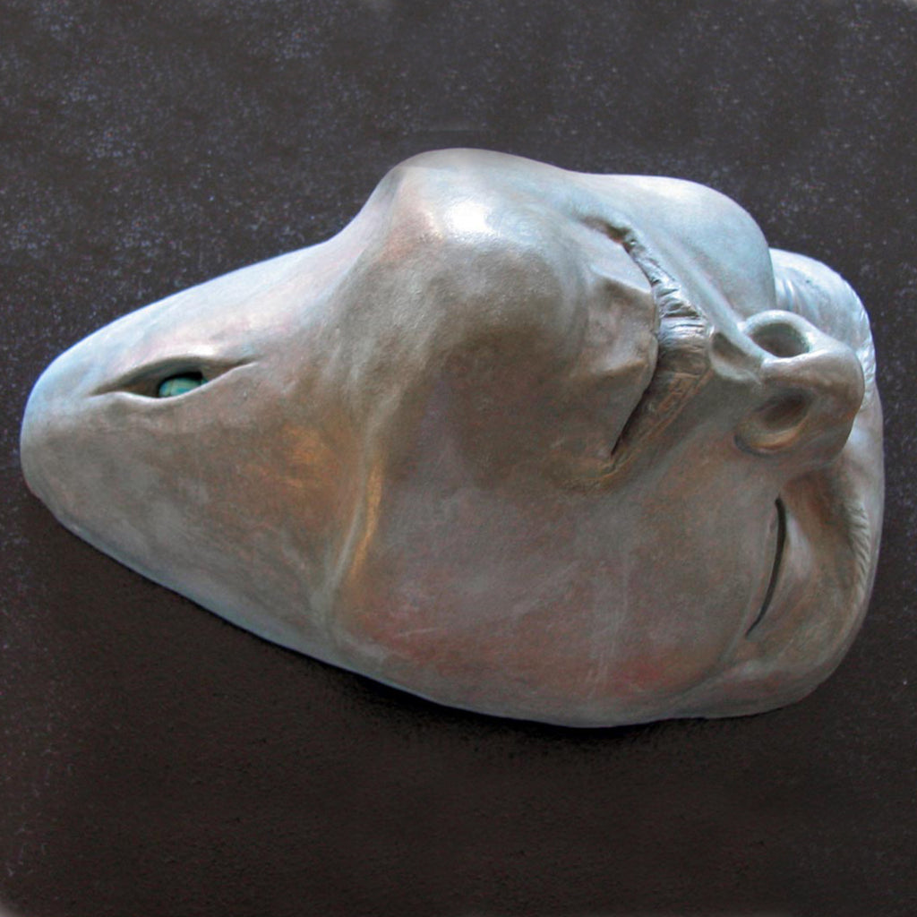 The artwork 'LUCKY' by Nora Chavooshian features a sculptural cast forton mask with bronze patina, displaying smooth, curved surfaces and ethereal qualities. The mask exhibits a serene expression with defined contours, subtle hollows, and a metallic silvery-gray finish that catches light, creating depth and dimension on its 42-inch form.