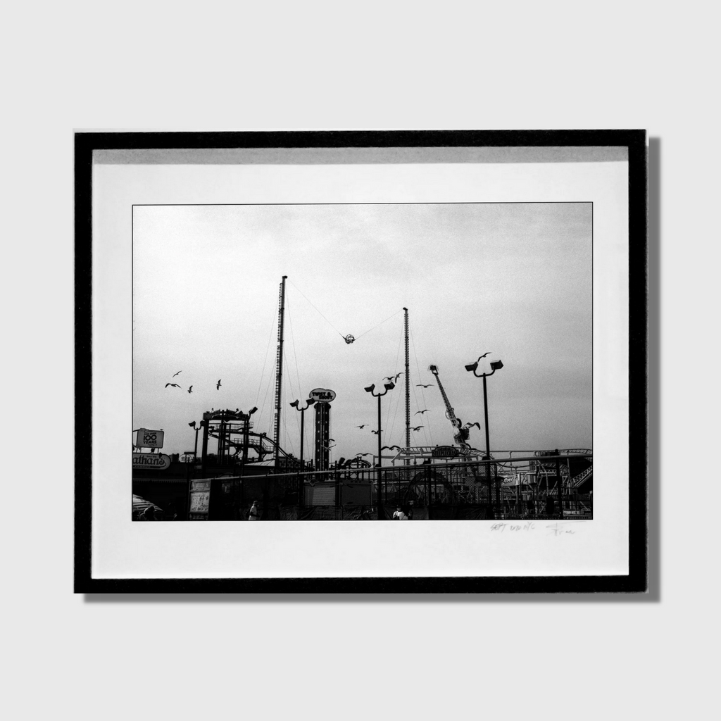 The artwork 'Luna Park' by Mari Sarai captures a monochromatic urban landscape featuring silhouettes of amusement park structures against an overcast sky. The black and white photograph showcases towering poles, light fixtures, and mechanical rides in stark contrast, creating a moody, atmospheric composition. Professionally framed in black, measuring 40x32 inches.