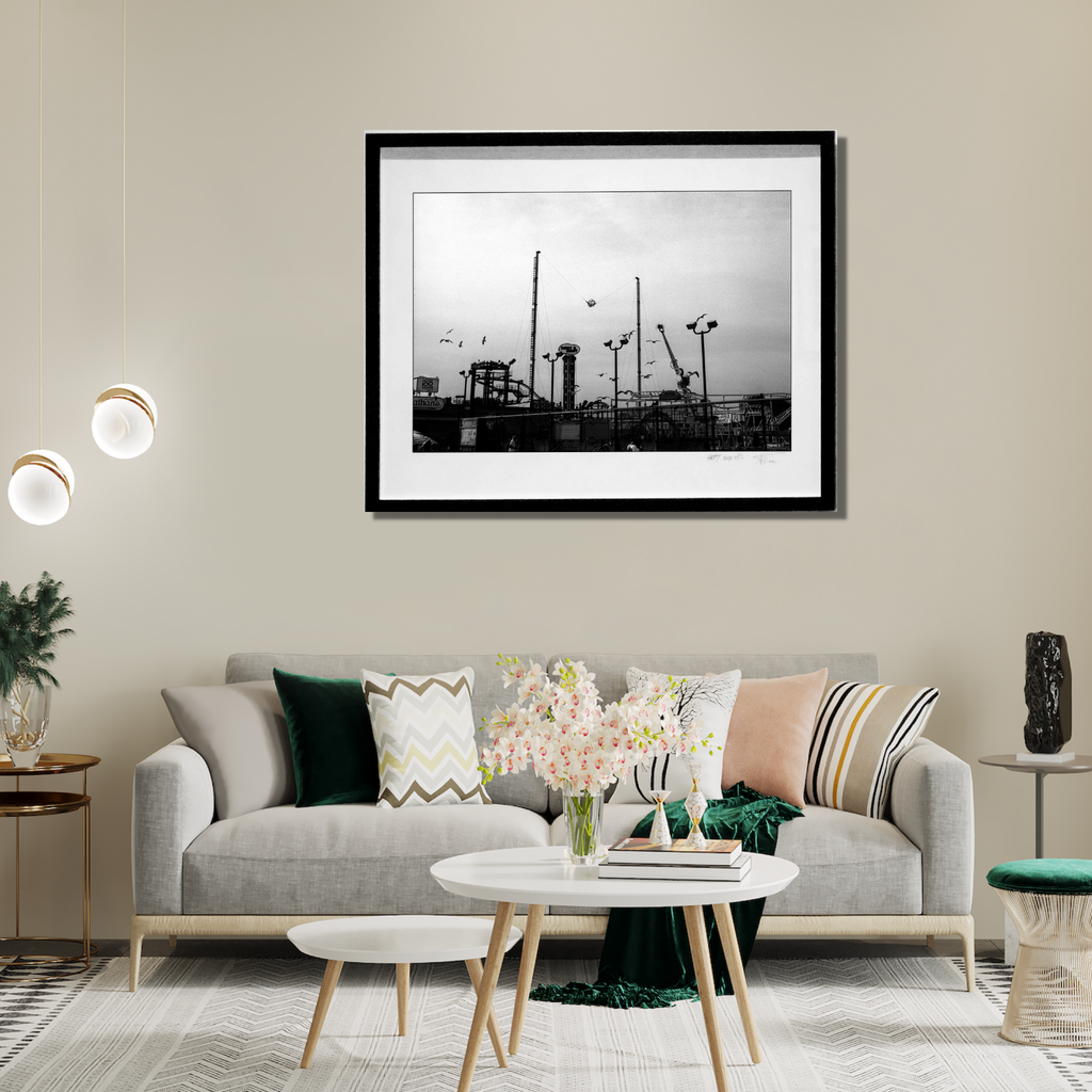 The artwork 'Luna Park' by Mari Sarai features a black and white photograph of an amusement park silhouette against a moody sky. Towering mechanical structures, poles, and carnival rides create a dramatic industrial skyline. The monochromatic composition is elegantly framed in black, capturing the nostalgic essence of a vintage fairground. - By Mari Sarai