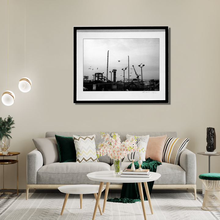 The artwork 'Luna Park' by Mari Sarai features a black and white photograph of an amusement park silhouette against a moody sky. Towering mechanical structures, poles, and carnival rides create a dramatic industrial skyline. The monochromatic composition is elegantly framed in black, capturing the nostalgic essence of a vintage fairground. - By Mari Sarai
