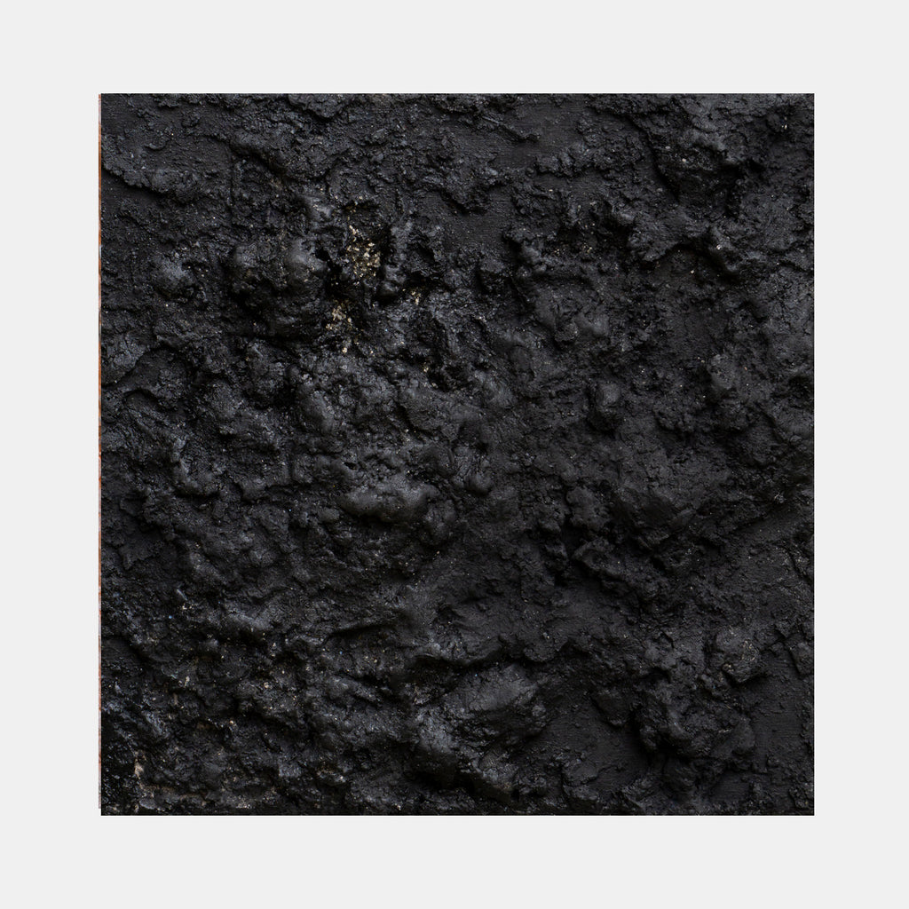 The artwork 'Magma' by Jacqueline Ferrante features a richly textured black surface resembling volcanic rock, with deep crevices and raised areas creating a dramatic topographical effect. Metallic mica flecks catch light against the dark plaster and charcoal backdrop, suggesting cooling lava. The mixed media piece combines smooth and rough textures in a monochromatic composition.