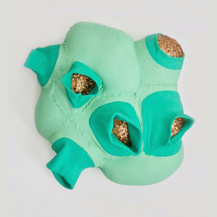 The artwork 'Marrow' by Hanna Washburn features an abstract sculptural form in mint green and turquoise hues. Created from recycled sweater cuffs, the piece reveals organic pod-like cavities filled with shimmering gold mica flakes. The textured surface shows intricate stitching patterns mounted on a wood panel, combining soft textile elements with metallic accents.