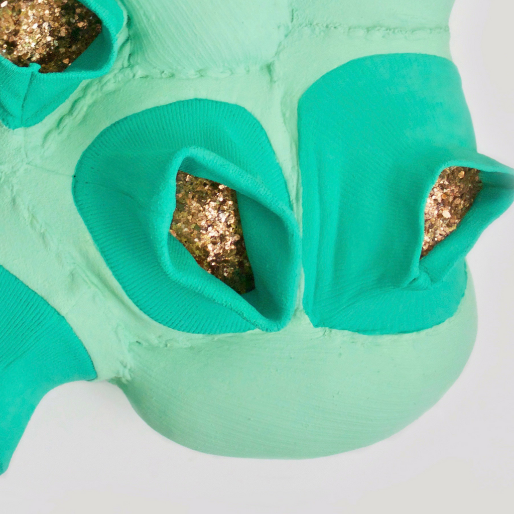 The artwork 'Marrow' by Hanna Washburn features an abstract sculptural form in mint green and turquoise hues. Created from recycled sweater cuffs, the piece reveals organic pod-like cavities filled with shimmering gold mica flakes. The textured surface shows intricate stitching patterns mounted on a wood panel, combining soft textile elements with metallic accents.