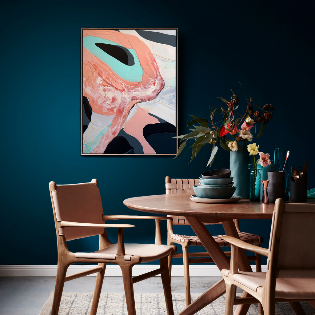 The artwork 'Meditation XXXII' by Renee Phillips features flowing abstract forms in coral pink and mint turquoise against a stark white background, creating a dynamic composition of organic shapes. The latex and spray paint on wood panel creates bold contrasts and smooth transitions, displayed in a modern dining space against a deep teal wall. - Renee Phillips