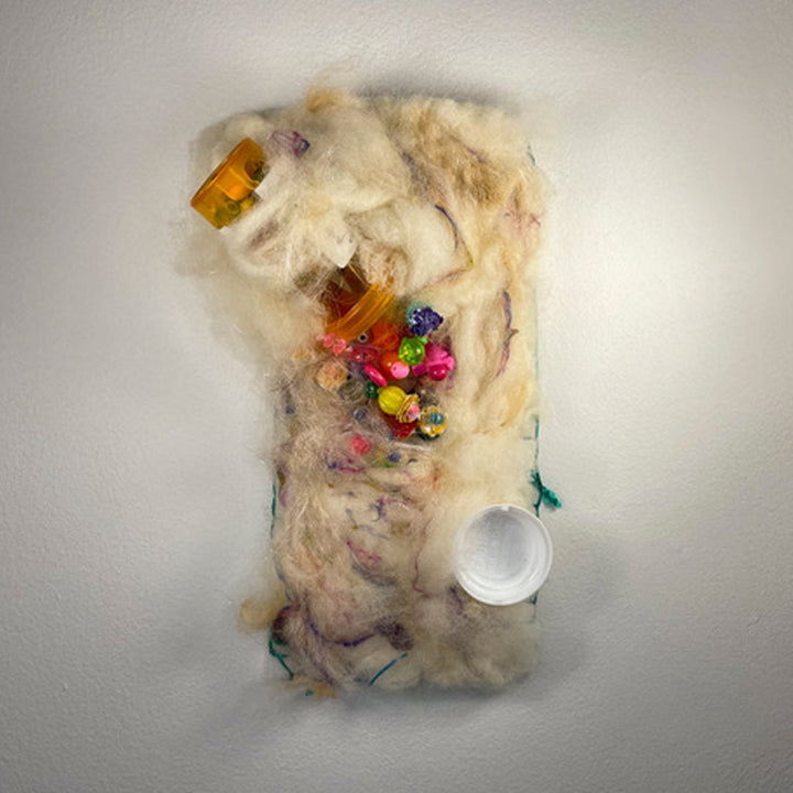 Miniature X by María Antonia Villaseñor-Marchal: Abstract textile sculpture featuring cream-colored wool and alpaca fibers with orange prescription bottles, vibrant multicolored beads, and sequins arranged organically. Mixed media piece incorporates silk thread, glass elements, and a white pill container against a soft textured background.