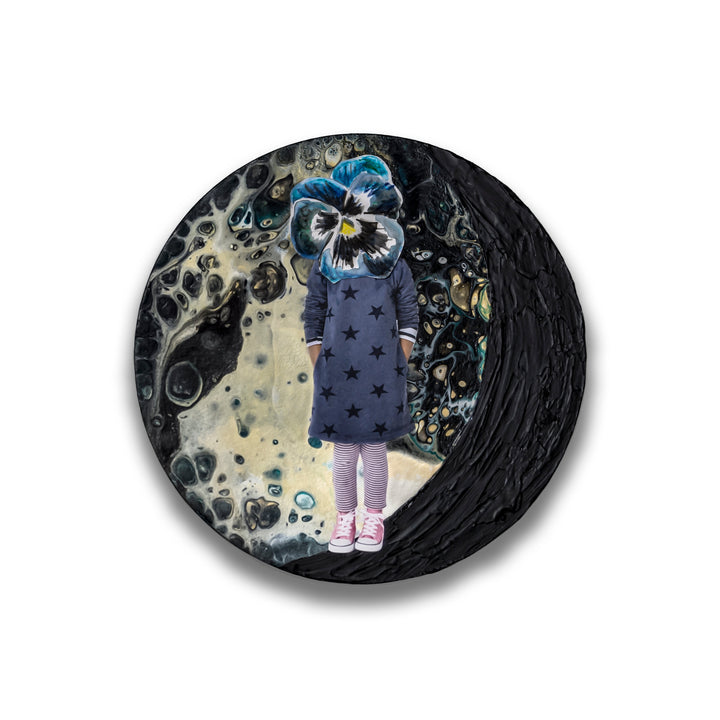 The artwork 'Moonchild' by Mary Younkin features a whimsical mixed-media composition on a circular wood panel. A figure in a star-patterned navy dress stands against a cosmic backdrop of swirling blacks, grays, and creamy whites. A vibrant blue pansy bloom serves as the head, creating a surreal collage effect. Textured acrylic creates a lunar, otherworldly atmosphere.