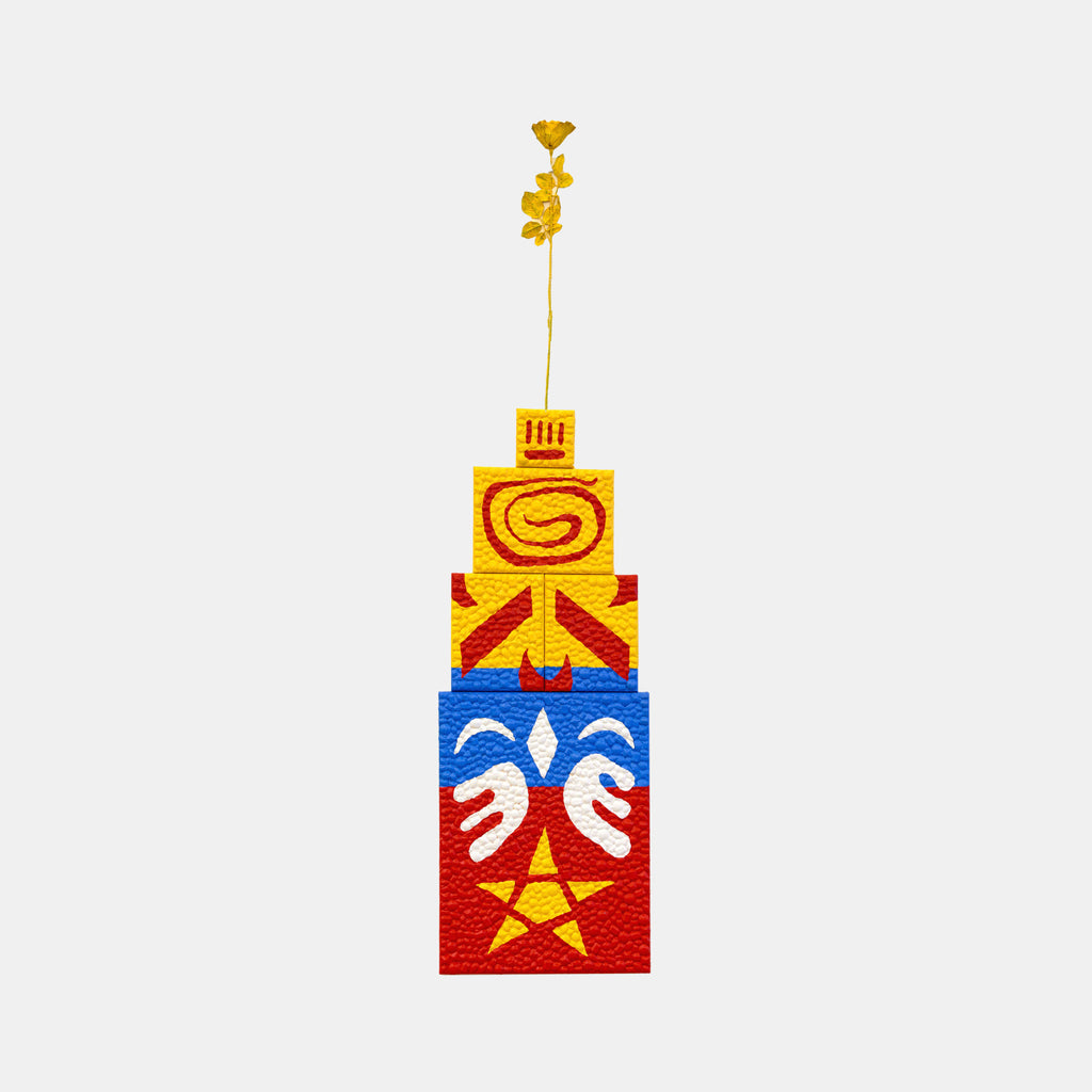 The artwork 'Neither Here Nor There D' by Marco DaSilva features a vibrant totem-like structure with stacked geometric blocks in yellow, red, and blue. Abstract symbols include a spiral motif, arrow patterns, and wing-like shapes, adorned with rhinestone accents. The composition rises vertically on white canvas, finished with polyurethane sheen. By Marco DaSilva.