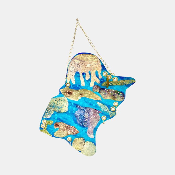 The artwork 'Ocean 2' by Sunny Chapman features vibrant abstract oceanscape in acrylic on paper, showcasing fluid turquoise blue waters intermingled with metallic gold accents and textured patches. The irregular-shaped piece hangs from a delicate gold chain, creating a dynamic wall art that captures the ethereal movement and shimmer of ocean waters.