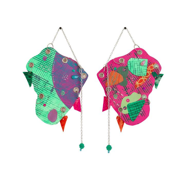 The artwork 'One Way Or Another' features two mirrored abstract shapes in vibrant pink and green, adorned with geometric patterns, metallic chains, and decorative elements. Mixed media piece combines glitter, sequins, plastic jewels, and handmade charms creating a playful butterfly-like composition. By Sunny Chapman