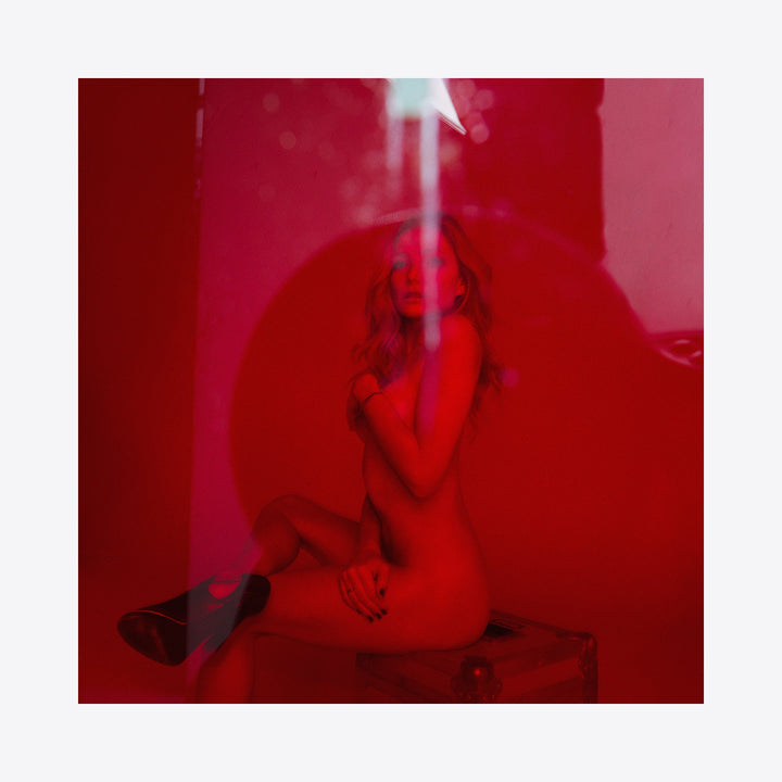 The artwork 'Clara in the Red Light' by Mari Sarai features a silhouette bathed in intense crimson lighting. A dreamy, atmospheric composition captures a figure seated on a geometric form, with ethereal smoke or mist rising upward. The archival pigment print creates a striking monochromatic effect in deep red tones, measuring 11x14 inches.