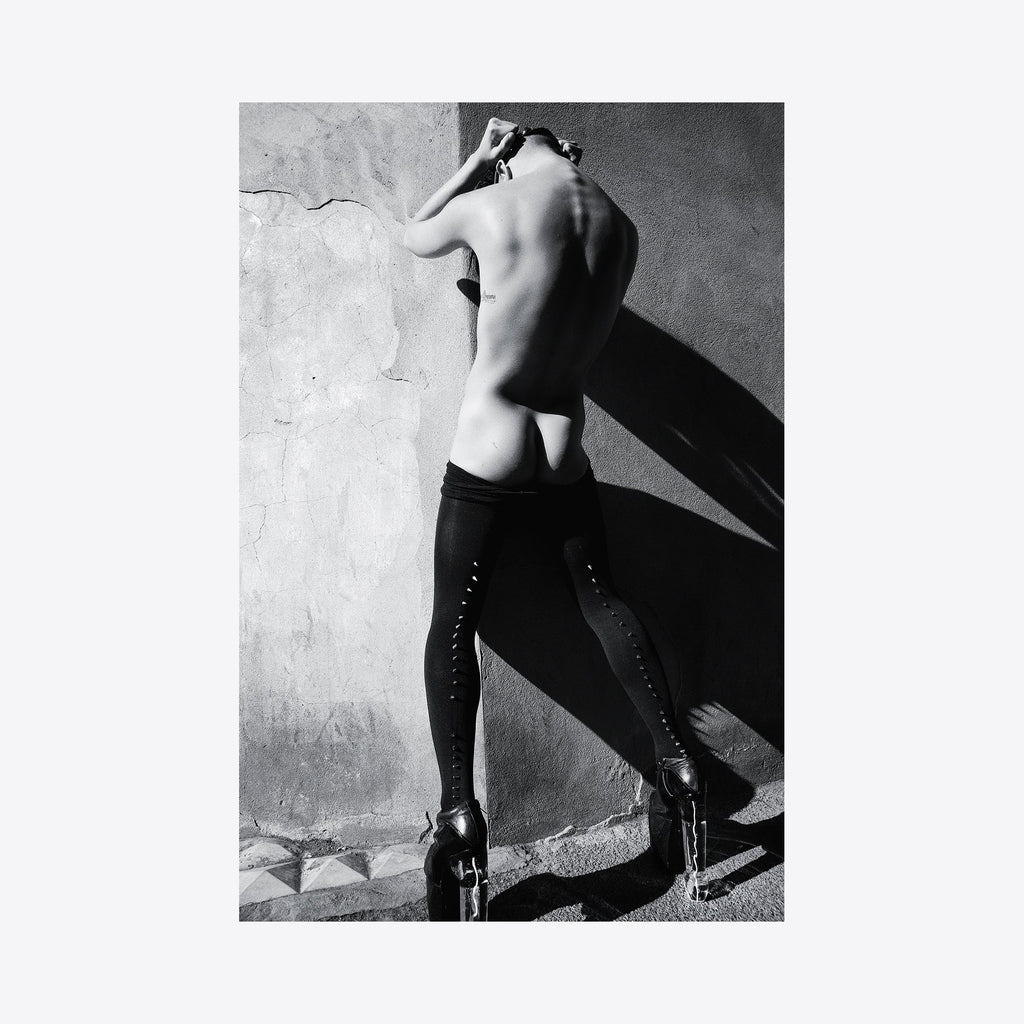 The artwork 'Cock Heel' by Mari Sarai features a dramatic black and white photograph showing a figure in knee-high laced boots against a textured concrete wall. Strong shadows and highlights create compelling contrast, while the composition emphasizes geometric angles and negative space. Archival pigment print, 11x14 inches.