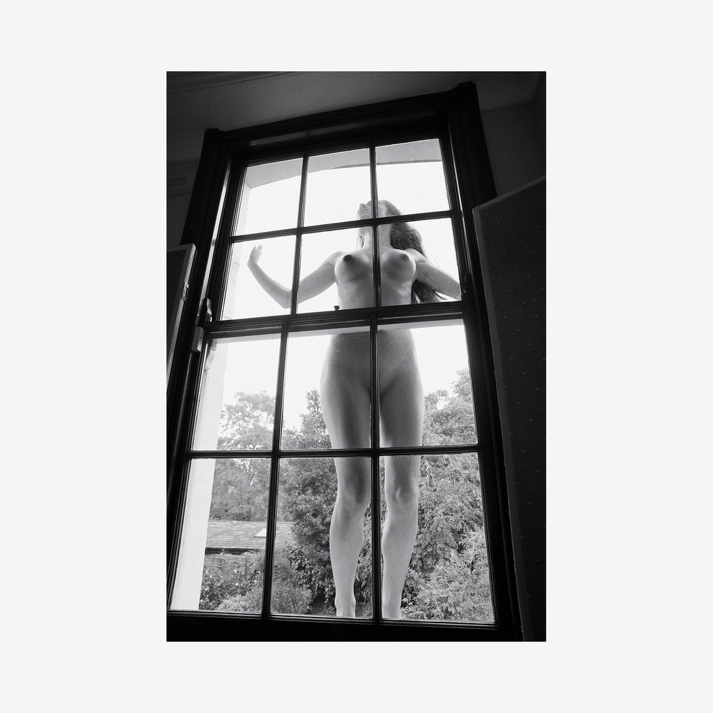 The artwork 'Girl in the Wonderland' is a dramatic black and white archival pigment print featuring a silhouette through a traditional paned window. The composition emphasizes geometric lines of the window frame against soft natural light, creating a dreamy, ethereal atmosphere. By Mari Sarai.