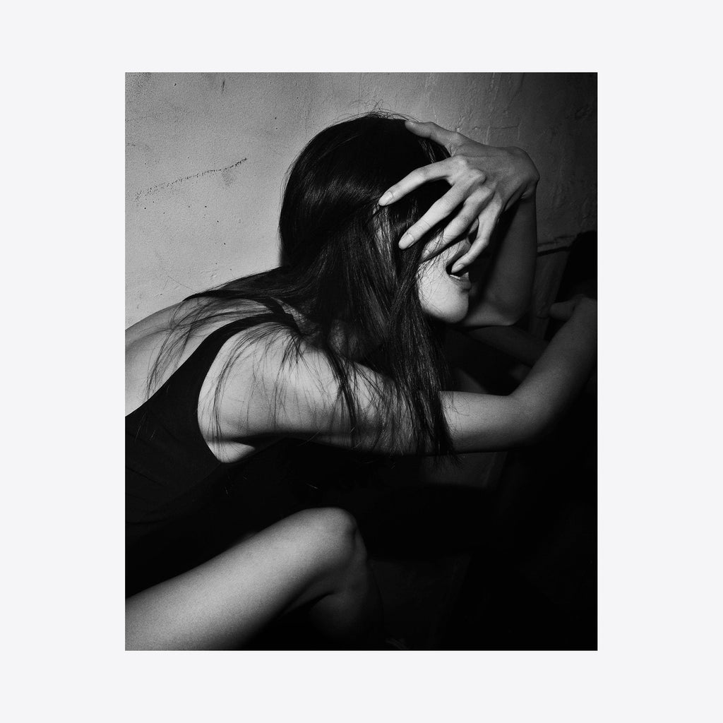 The artwork 'Hand on Face' features a dramatic black and white composition showing long dark hair cascading over shoulders, with an elegantly positioned hand creating a striking gesture against a textured background. The archival pigment print captures intimate shadows and light contrasts in a minimalist style. By Mari Sarai.