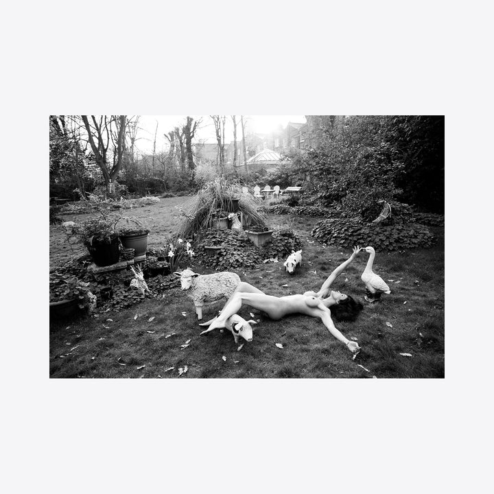 The artwork 'Janice in Wonderland' captures a dreamy scene in a garden setting, rendered in black and white. A figure reclines gracefully on dark earth, surrounded by cats in a whimsical arrangement. The composition features bare trees, garden debris, and rustic elements including planters and natural foliage, creating an ethereal, fairy tale atmosphere. Archival pigment print by Mari Sarai.