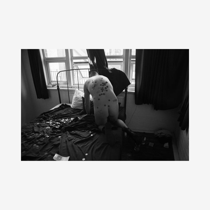 The artwork 'Money Bed' by Mari Sarai: A dramatic black and white archival pigment print capturing a minimalist bedroom scene with scattered items on dark bedding near a window with dark curtains. The composition creates a stark contrast between light and shadow, emphasizing geometric shapes and textures in the 11x14 inch frame by Mari Sarai.