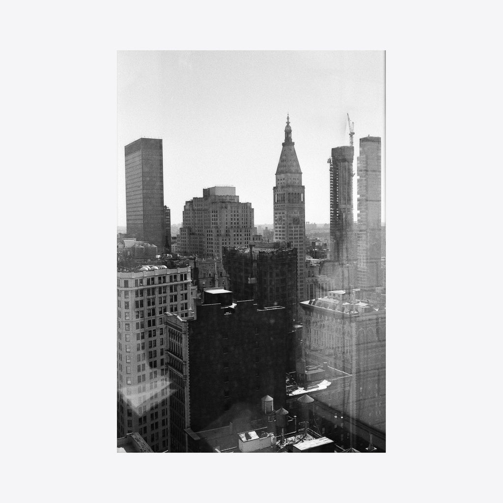 The artwork 'NY Melancholy' captures a moody black and white urban landscape of Manhattan's iconic skyline, featuring towering skyscrapers including the distinctive Met Life Building. The archival pigment print presents a dramatic contrast between light and shadow, showcasing architectural details against a misty cityscape by Mari Sarai.