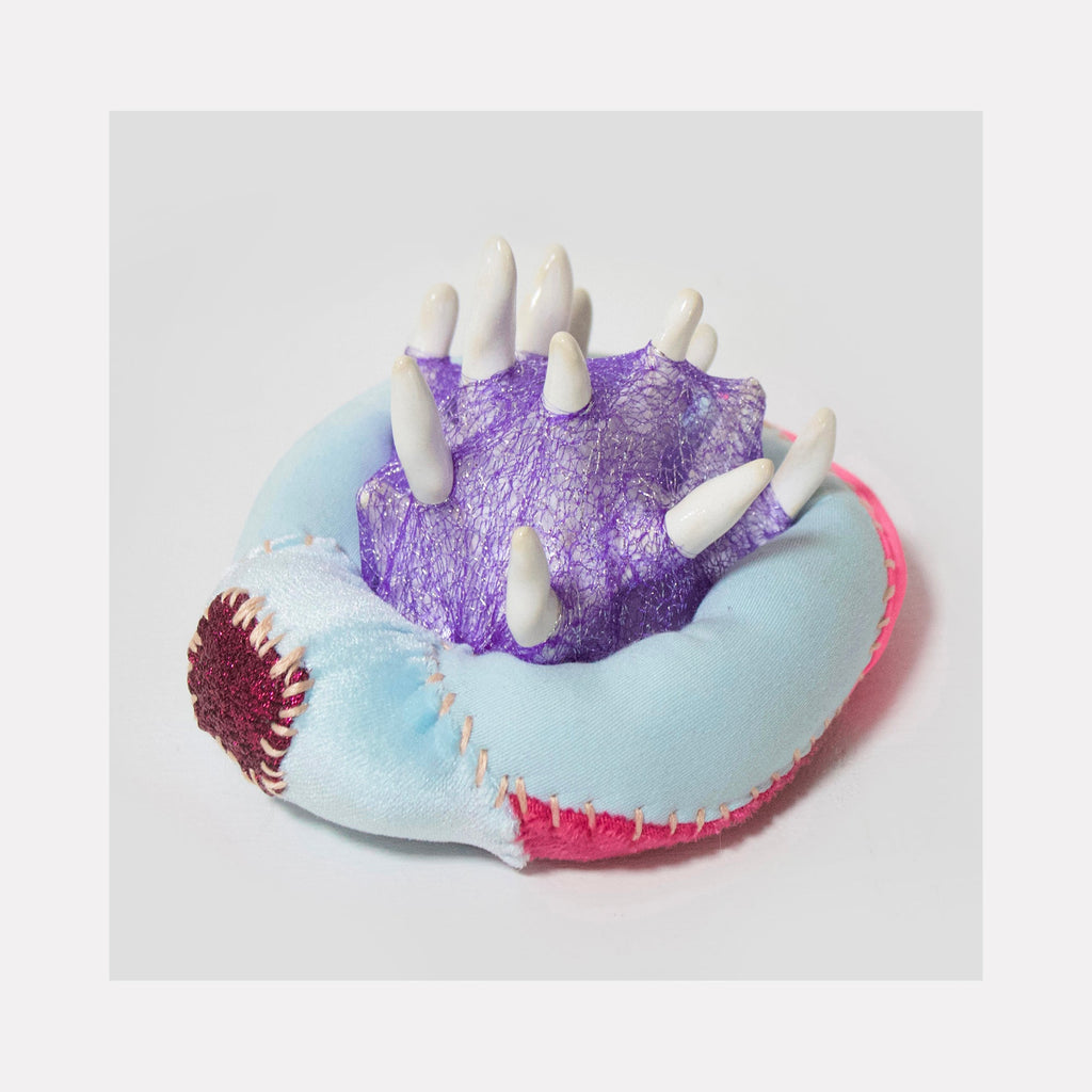 The artwork 'Pedicellaria' by Hanna Washburn features a whimsical mixed-media sculpture combining glazed ceramic spikes emerging from a purple textured surface, set atop a soft pastel blue and pink cushion-like base with visible decorative stitching and burgundy fabric accent detail. The piece harmoniously blends hard and soft materials.