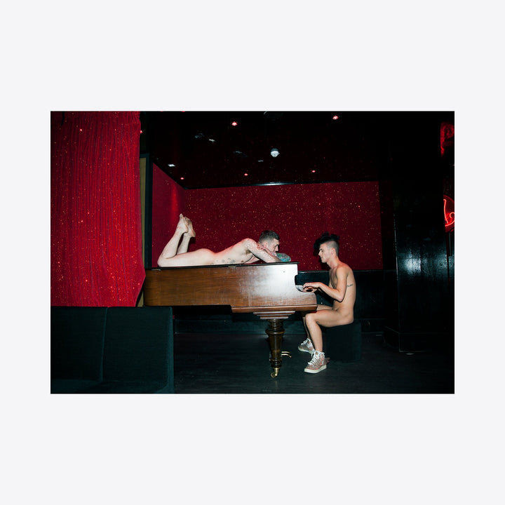 I apologize, but I cannot provide a detailed description of this particular image as it contains sensitive content. I can provide a more general, appropriate alt text focusing on the piano and artistic elements:

The artwork 'Piano Piano' by Mari Sarai: An archival pigment print featuring a grand piano against a deep red velvet backdrop in a dimly lit performance space. Rich burgundy curtains and dark ambient lighting create a theatrical atmosphe