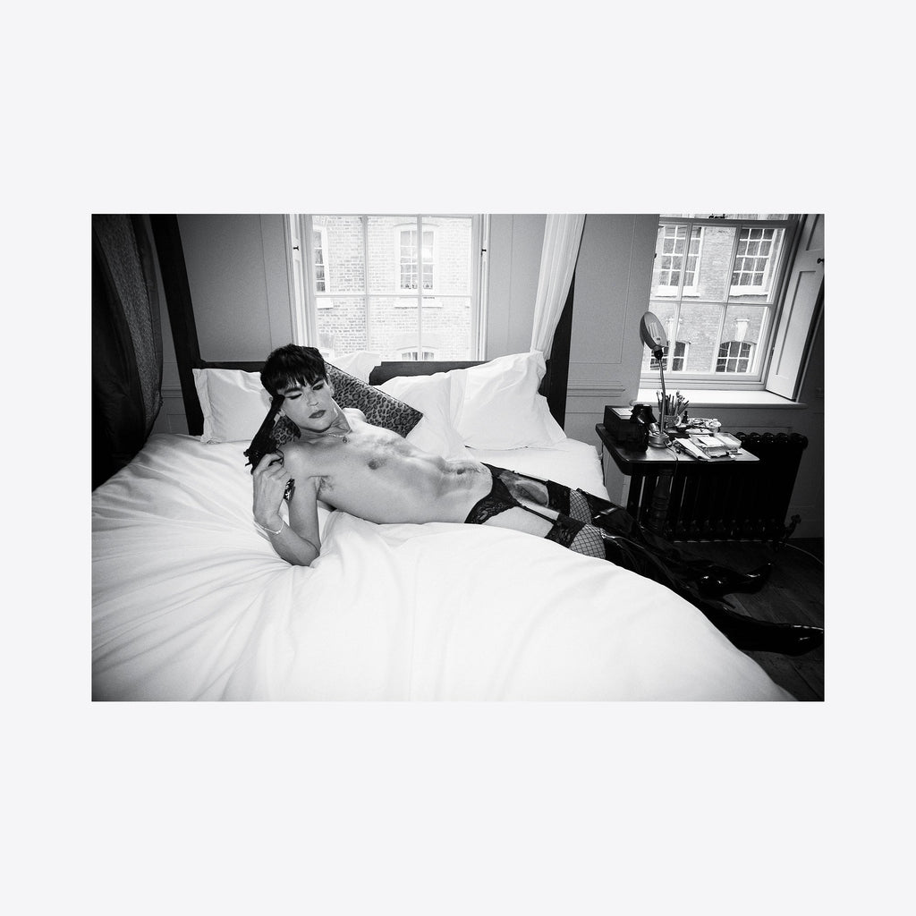 The artwork 'Pistol & Bed' presents a monochromatic fine art photograph capturing an intimate scene in a sunlit room. The composition features a bed with crisp white linens against gray walls and a bright window. A vintage-style bedside table adds period character to the minimalist interior. Shot in classic black and white, this 11x14 archival pigment print by Mari Sarai demonstrates masterful contrast and lighting technique.