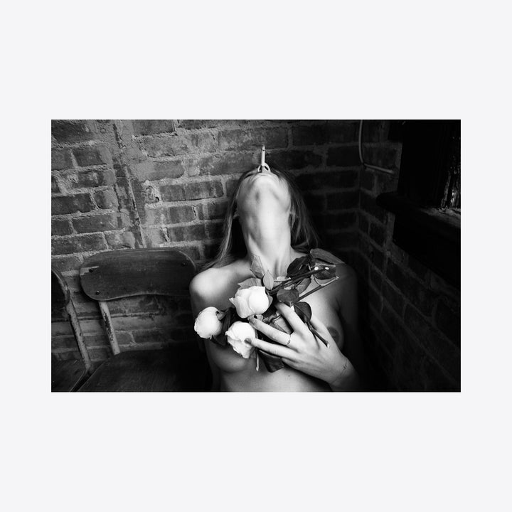 The artwork 'Rose & Cigarette' by Mari Sarai captures a dramatic black and white composition against a textured brick wall backdrop. White roses contrast with shadows, creating a moody aesthetic. The monochromatic archival pigment print emphasizes light and shadow interplay, displaying artistic mastery in film photography. 24x20 inches.