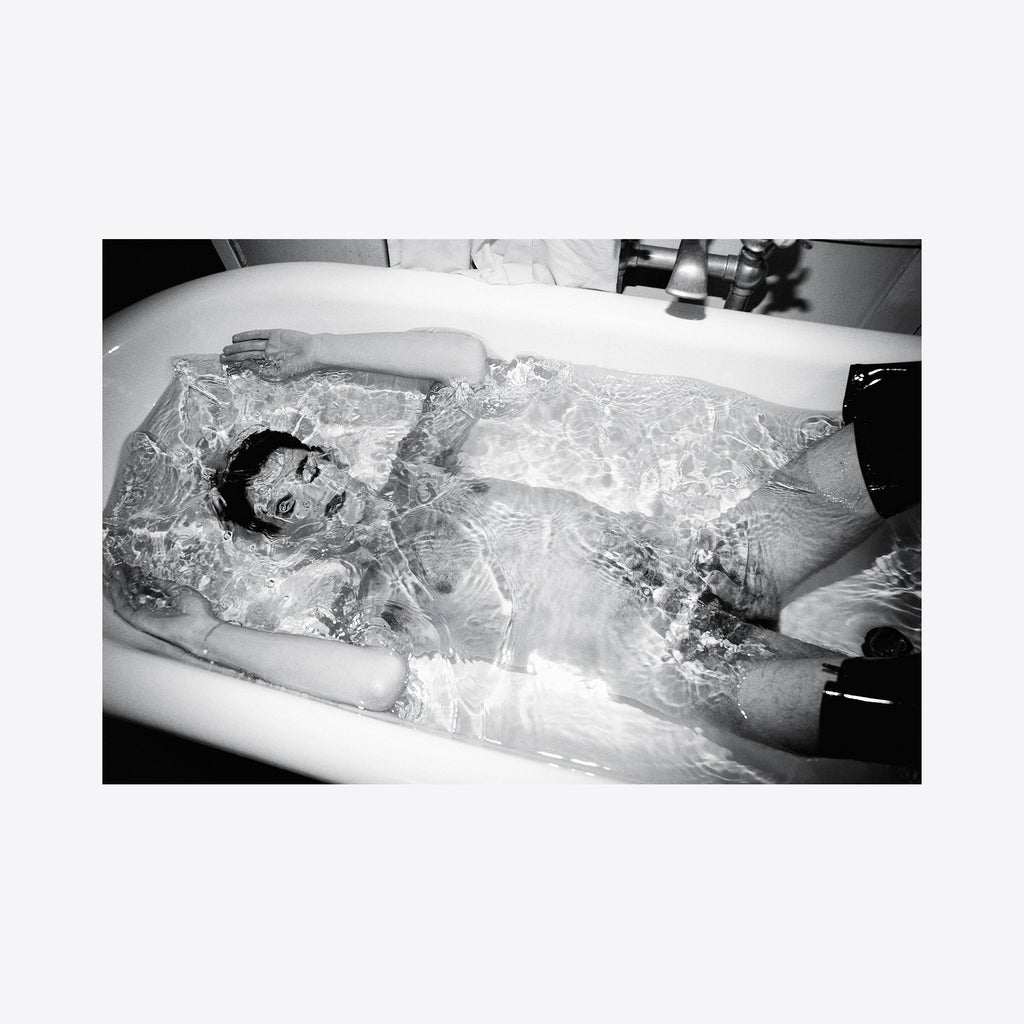 The artwork 'Simulated Experience' features a monochromatic bathtub scene captured in stark black and white photography. Rippling water creates intricate patterns of light and shadow across a figure submerged in a porcelain tub, creating an ethereal atmospheric effect. The archival pigment print showcases dramatic contrast and textural elements. By Mari Sarai.