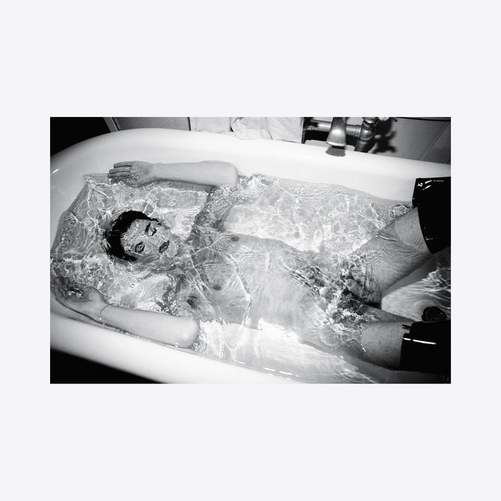 The artwork 'Simulated Experience' features a monochromatic bathtub scene captured in stark black and white photography. Rippling water creates intricate patterns of light and shadow across a figure submerged in a porcelain tub, creating an ethereal atmospheric effect. The archival pigment print showcases dramatic contrast and textural elements. By Mari Sarai.