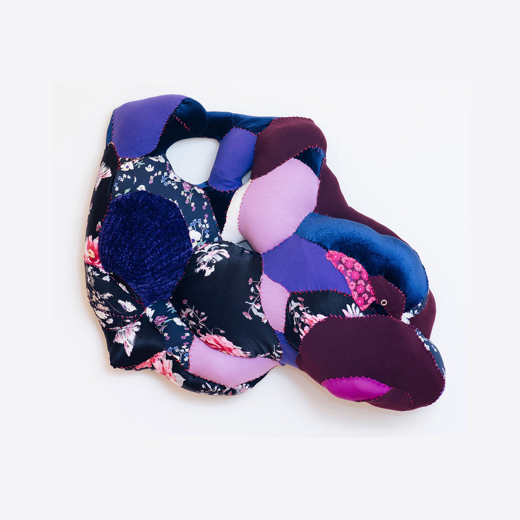 The artwork 'Supine Twist' by Hanna Washburn features an abstract textile composition of curved, interwoven shapes in deep purple, royal blue, and soft pink. The piece combines floral printed fabric with solid colored recycled materials, creating a dimensional sculptural effect. Stitched details and quilted textures add depth to this wall-mounted textile artwork.