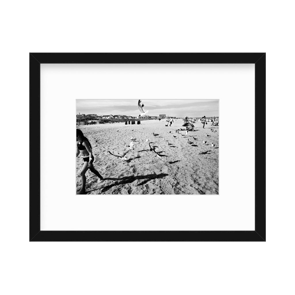 The artwork 'Coney Island' by Mari Sarai captures a dynamic black and white beach scene, with seagulls taking flight across a sandy expanse. Figures are scattered across the shoreline, creating dramatic shadows in the bright sunlight. The composition features a stark contrast between light sand and dark silhouettes, framed in a classic black frame with white matting. Limited edition photograph #20, by Mari Sarai.