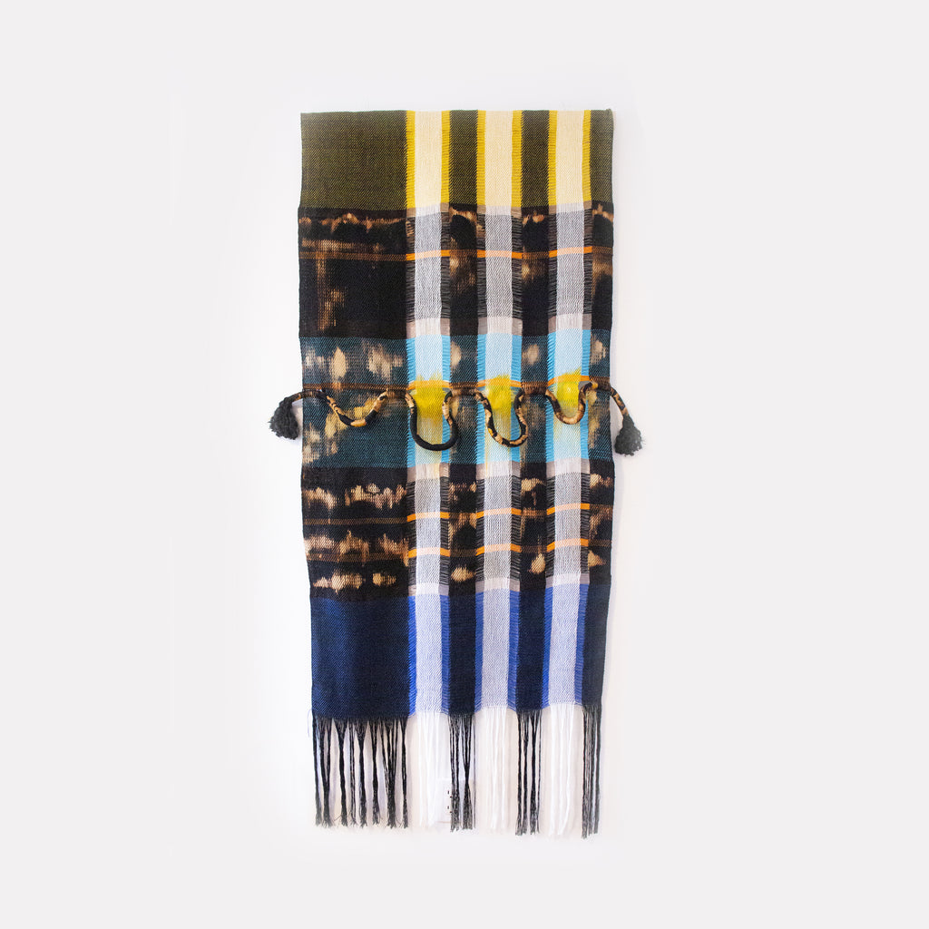 The artwork 'Fragments Of The Sky' by Kathie Halfin features vertical stripes in yellow, blue, and navy, with tie-dye-like brown bands creating a horizon effect. The textile piece incorporates copper wire elements and hanging fringe details at the bottom, crafted from cotton and linen. The composition creates an abstract interpretation of a skyscape, with contrasting light and dark elements.