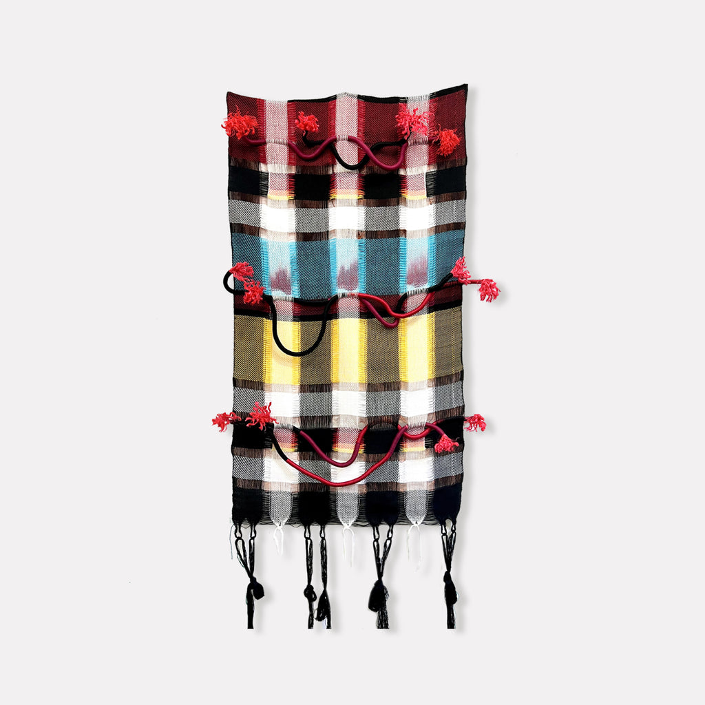 The artwork 'Resisting The Fire' by Kathie Halfin features a vertical textile composition with bold plaid patterns in red, turquoise, yellow, and black stripes. Woven cotton fabric is enhanced with decorative red tassels and black rope elements, creating dynamic textural contrasts. Copper wire accents curve through the piece, while linen details and hanging tassels complete the 60x35 inch wall hanging.