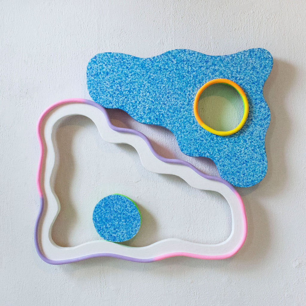 The artwork 'Floopy Float' by Ryan Patrick Martin features an abstract sculptural composition with a textured bright blue irregular shape containing a yellow-rimmed circular opening, paired with a curving white and pink bordered form. The wooden wall sculpture combines smooth and granular textures in playful geometric forms.