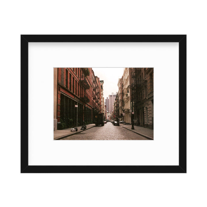 The artwork 'Ghost Neighborhood in Soho' by Mari Sarai captures a moody sepia-toned street scene in New York's historic Soho district. The composition features classic cast-iron buildings with fire escapes, a cobblestone street stretching into the distance, and vintage urban architecture. The black and white framed photograph creates a timeless, atmospheric perspective. By Mari Sarai.