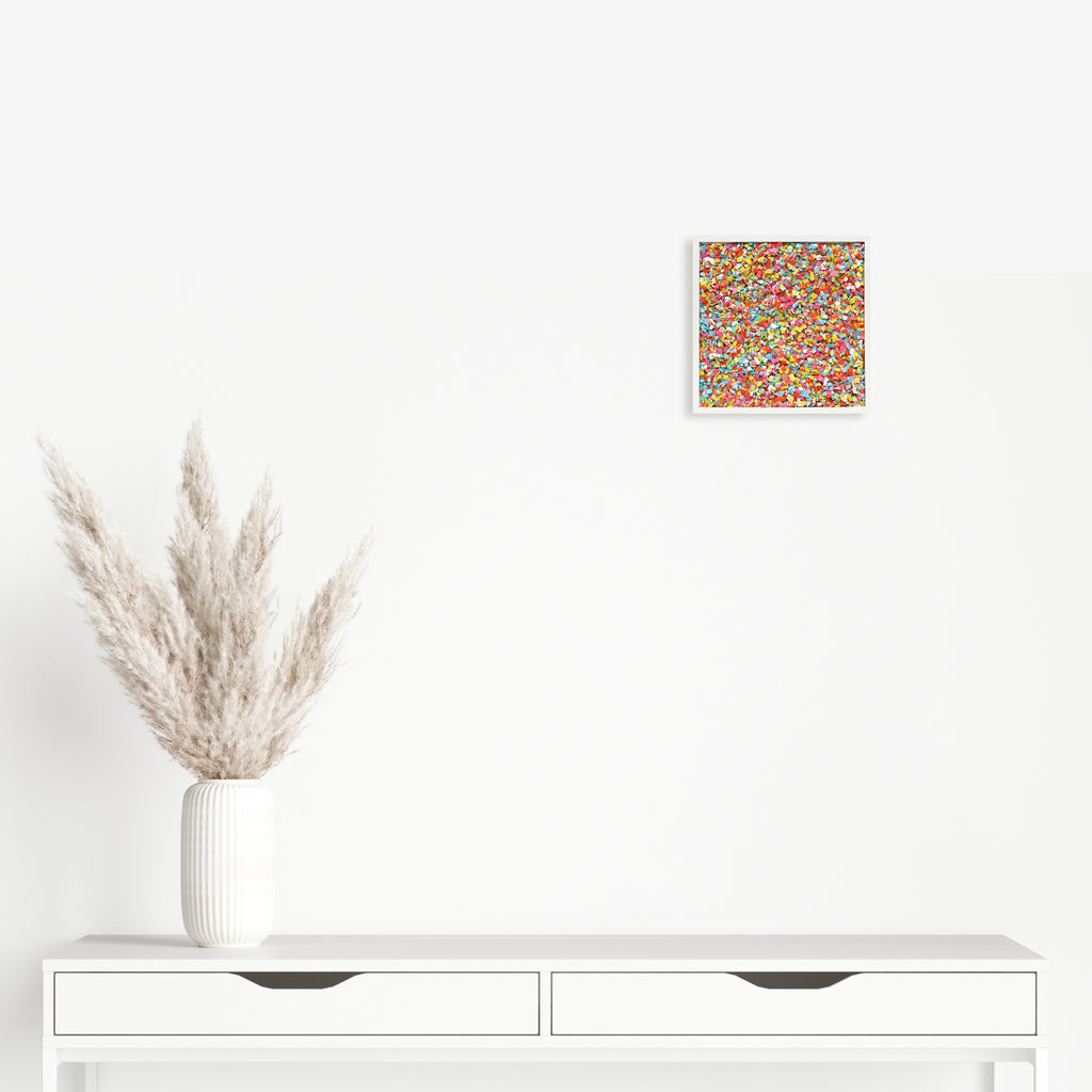 The artwork 'Party Popper' by Lori Kirkbride features a vibrant, confetti-like pattern of multicolored dots and specks scattered across a square panel. The acrylic and resin composition creates a festive, energetic texture reminiscent of scattered party confetti, with bright yellows, reds, blues, and greens dancing across the 8.25-inch surface.