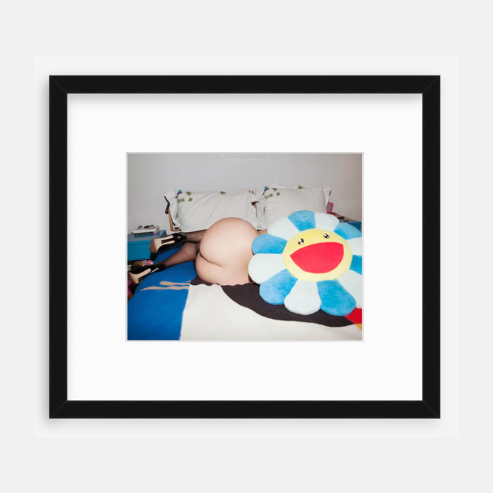 The artwork 'Peach Bum' by Mari Sarai showcases an artistic composition featuring a plush flower toy with bright blue petals and a cheerful yellow center, set against white bedding and blue fabric. The image is presented in a sleek black frame with white matting, displaying contemporary photography style.