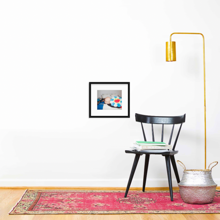 The artwork 'Peach Bum' by Mari Sarai, displayed in a modern interior setting with a black wooden chair, vintage pink rug, gold floor lamp, and silver woven basket. Framed in classic black, the 8x10 artist print creates a striking focal point against the clean white wall.