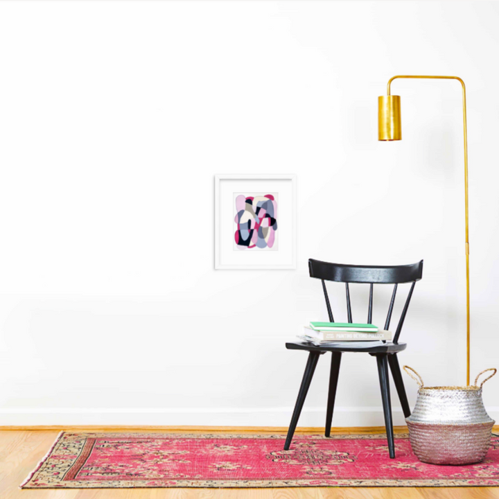 The artwork 'Pink and Grey on Sand' by Alicia Little features abstract organic shapes in soft pink and grey hues on a sandy background, displayed in a white frame against a minimalist white wall, accompanied by a black wooden chair, brass floor lamp, and vibrant pink vintage rug.