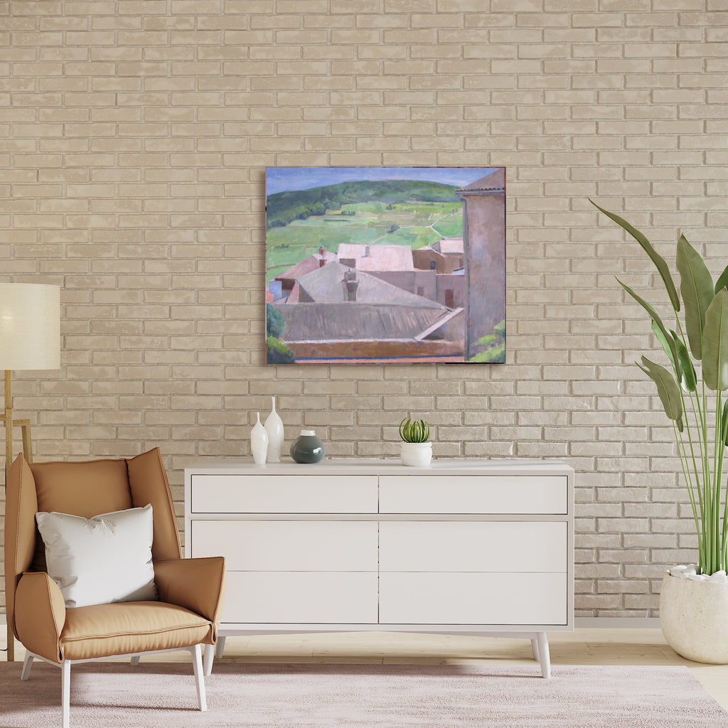 The artwork 'Place de Bretagne' by Peter Colquhoun depicts an elevated view of French village rooftops in muted tones. The oil on linen painting showcases terracotta-colored buildings against rolling green hills and a soft blue sky. The architectural forms are rendered in a contemporary impressionist style, capturing the rustic charm of Brittany's countryside. The composition is framed by distant mountains and features angular geometric shapes of